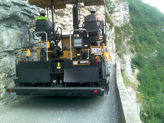 Асфальтоукладчик Cat AP655D MTS выполняет укладку покрытия на подъеме узкой извилистой дороги в горах Франции